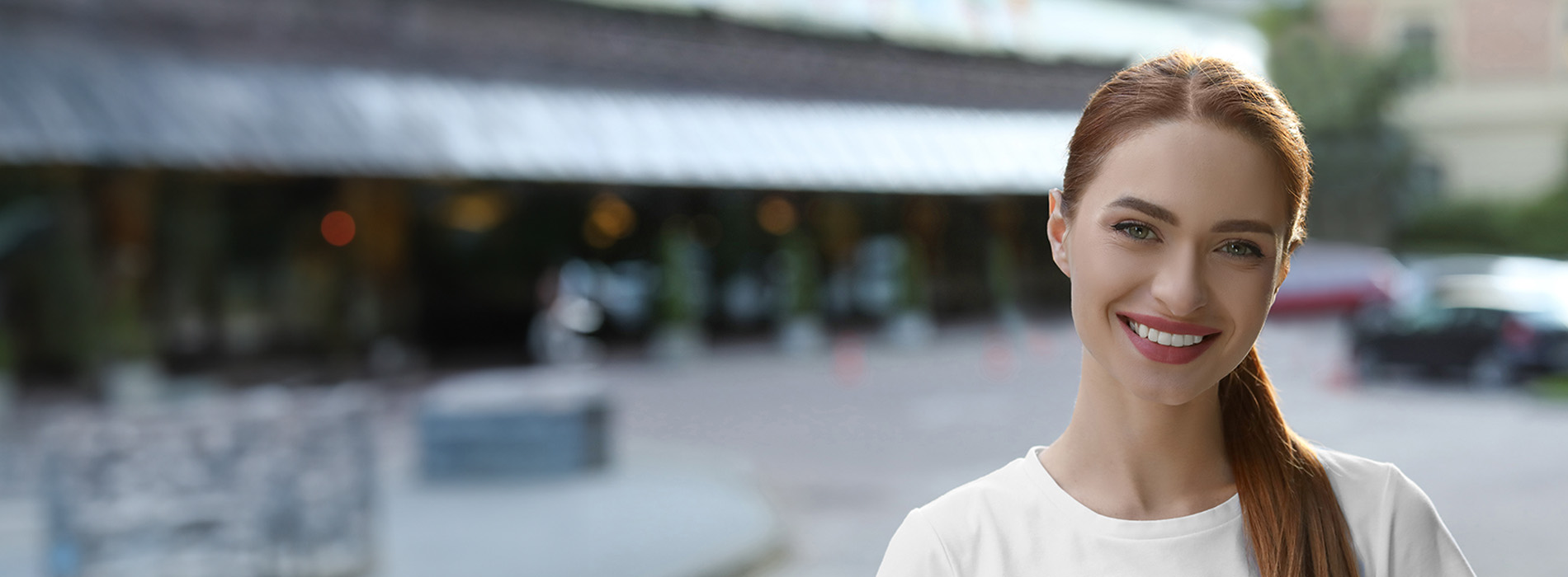 The image shows a woman with brown hair, wearing a white top, smiling at the camera. She is standing in front of a building with a sign that reads  BAR,  and there s a reflection of the building on the glass surface behind her.