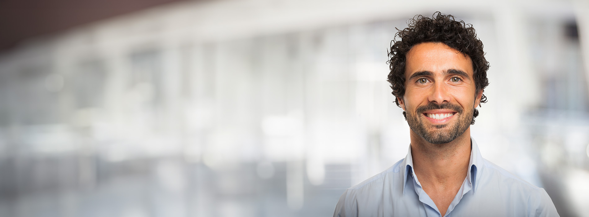 The image shows a man with dark hair and a beard, wearing a blue shirt and standing in front of a blurred background that suggests an interior space.