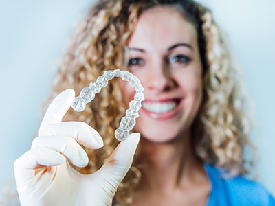 The image shows a person holding up a transparent dental retainer with their hand, while they are smiling.