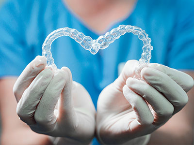 A clear dental retainer held by a pair of hands, with a blue background and a medical glove visible.