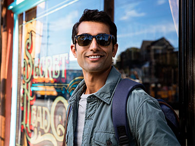 A young man in sunglasses, a backpack, and a jacket is standing outside a shop with a sign that reads  Sweet Treats.