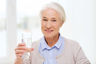 The image features an elderly woman holding a glass of water, smiling slightly and looking directly at the camera.