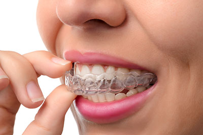 The image shows a person holding a toothbrush with a toothpaste strip on their finger, positioned in front of an open mouth displaying straight white teeth.