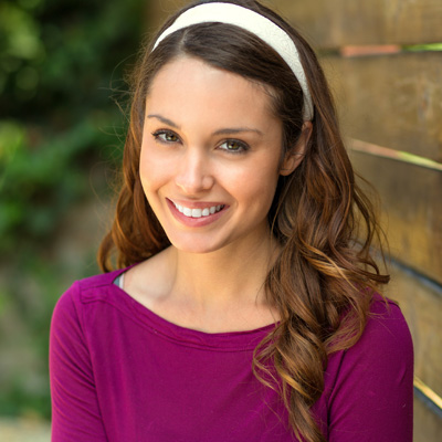 A smiling woman with long brown hair, wearing a purple top and a white headband, poses against a wooden fence.