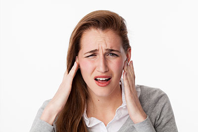 A woman in a grey blouse with her hand on her ear, appearing to be experiencing discomfort or pain.