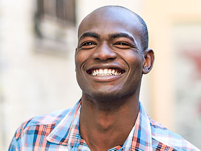 The image features a man with a broad smile, looking directly at the camera. He has short hair and is wearing a plaid shirt. There s a building with a balcony in the background.