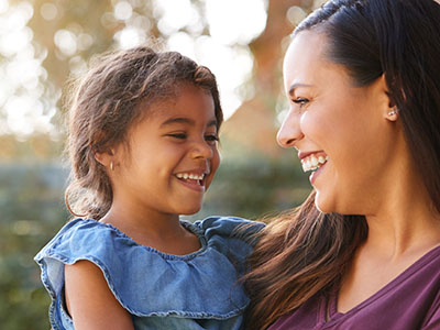 A woman and a child smiling, with the woman holding the child in an outdoor setting.