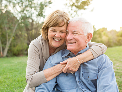 The image shows an elderly couple, a man and a woman, embracing each other outdoors. They are both smiling and appear to be in a relaxed, happy moment.