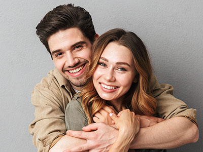 A young couple embracing in a warm, intimate pose, with the man on the left and woman on the right, both smiling and looking directly at the camera.