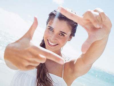 The image shows a woman taking a selfie with her hand, smiling at the camera while standing on a beach.