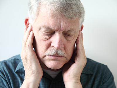 The image shows an older man with a mustache, wearing glasses and holding his face with both hands. He appears to be in a state of distress or discomfort.